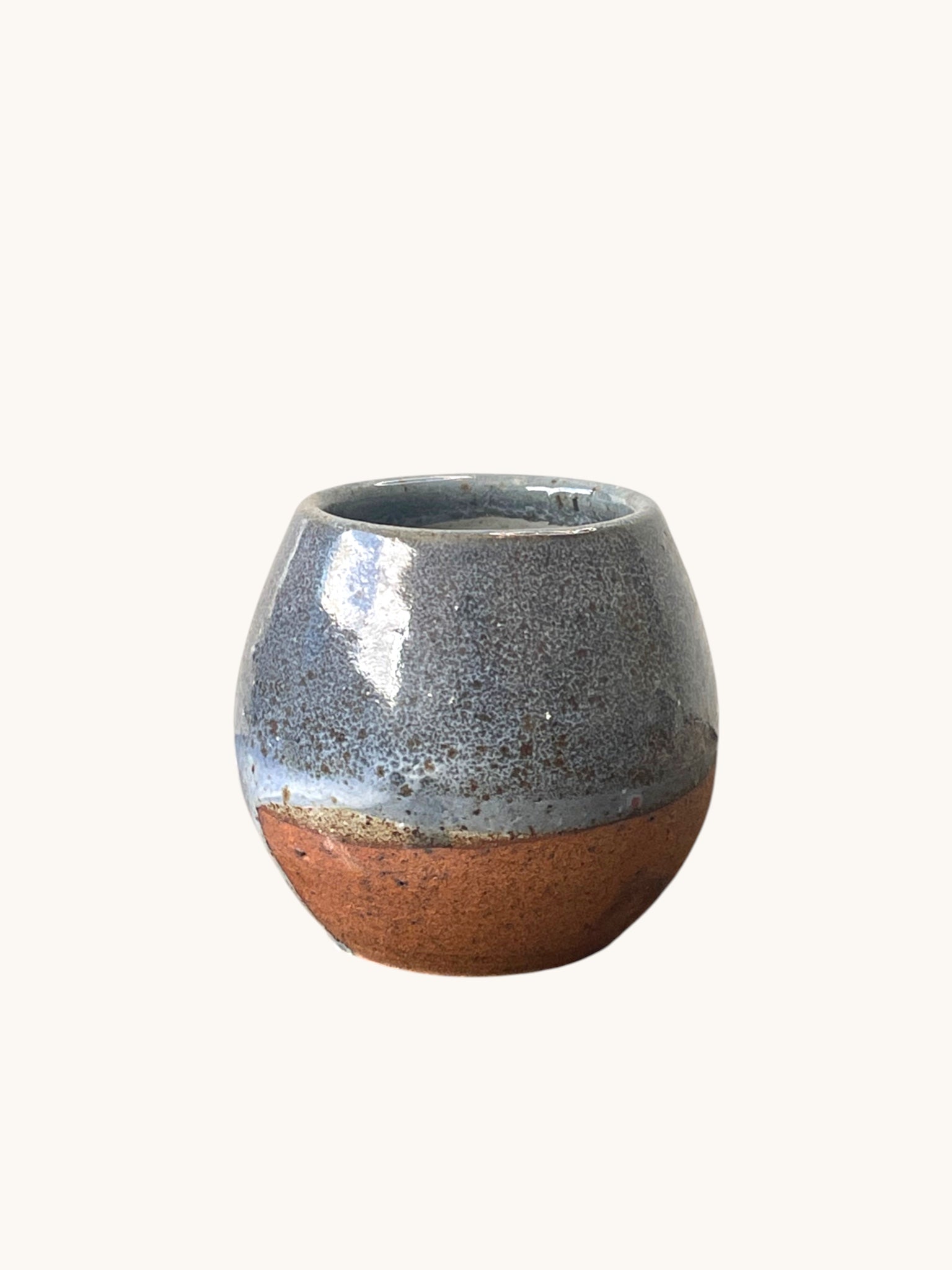 Matera Rob Original forma globular con vidriado gris acero brillante y base terracota natural, fondo crema. Matera Rob®
